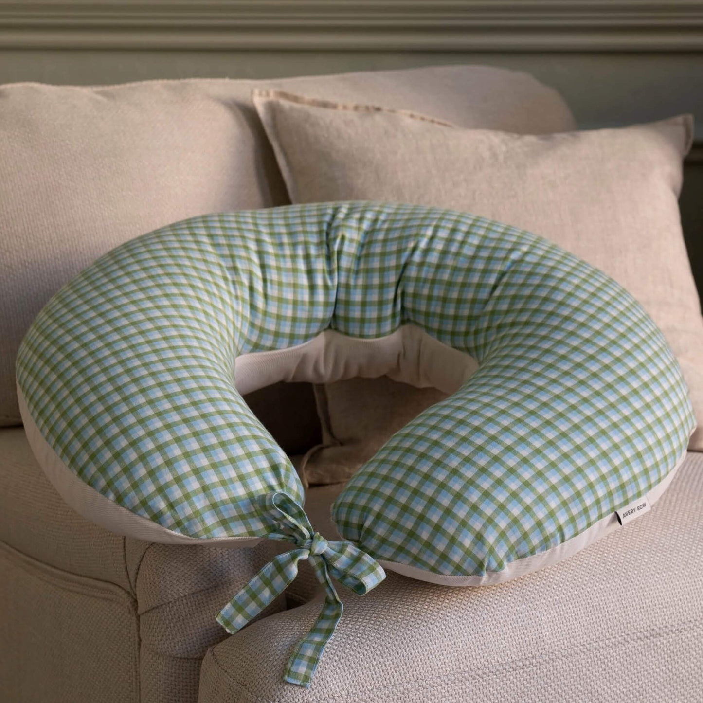 A U-shaped nursing pillow with a green and white gingham pattern, resting on a beige upholstered sofa with matching pillows.
