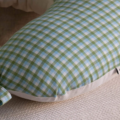 A close-up of a curved feeding pillow with a green, light blue, and white gingham plaid pattern, resting on a textured beige woven surface.