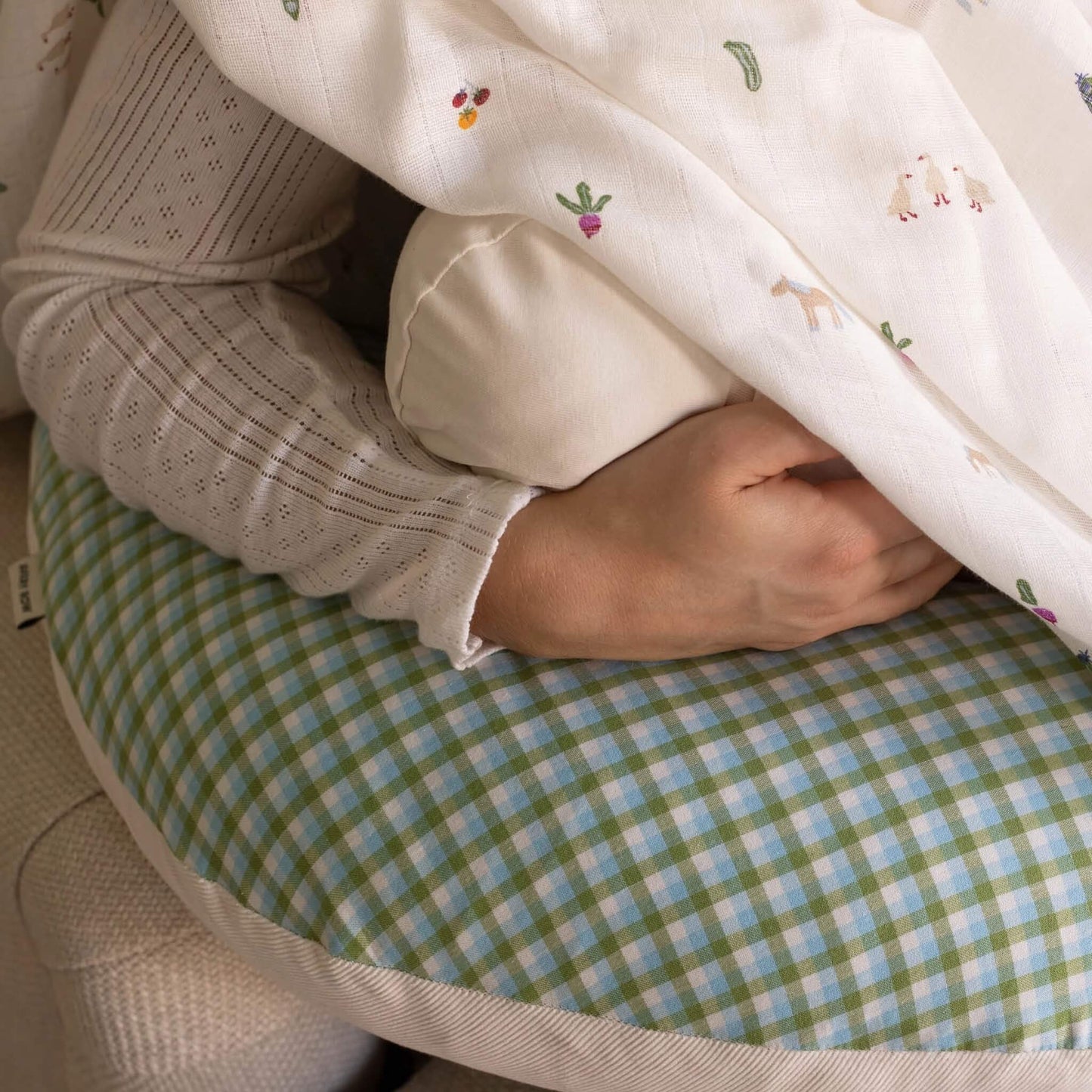 A close-up of a person cradling a baby support pillow. The pillow features a green and light blue gingham pattern with a cream-colored fabric border.