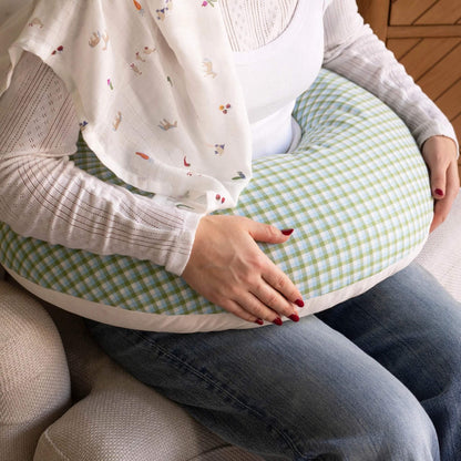A womansitting on a sofa holding a U-shaped nursing pillow. The pillow has a light green and white gingham pattern with a white fabric trim.