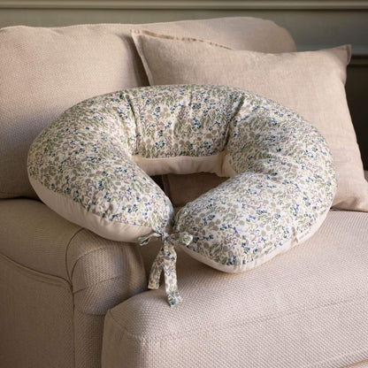 A U-shaped nursing pillow with a blue and green floral pattern on a cream background, resting on a beige fabric couch.