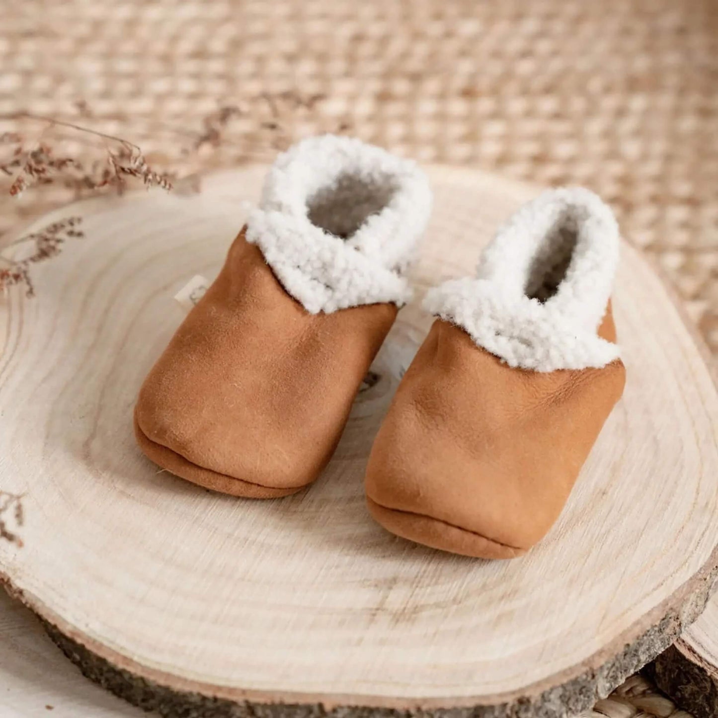 Baa Baby tan lambskin booties with soft wool trim, displayed on a natural wooden slice with a woven background.