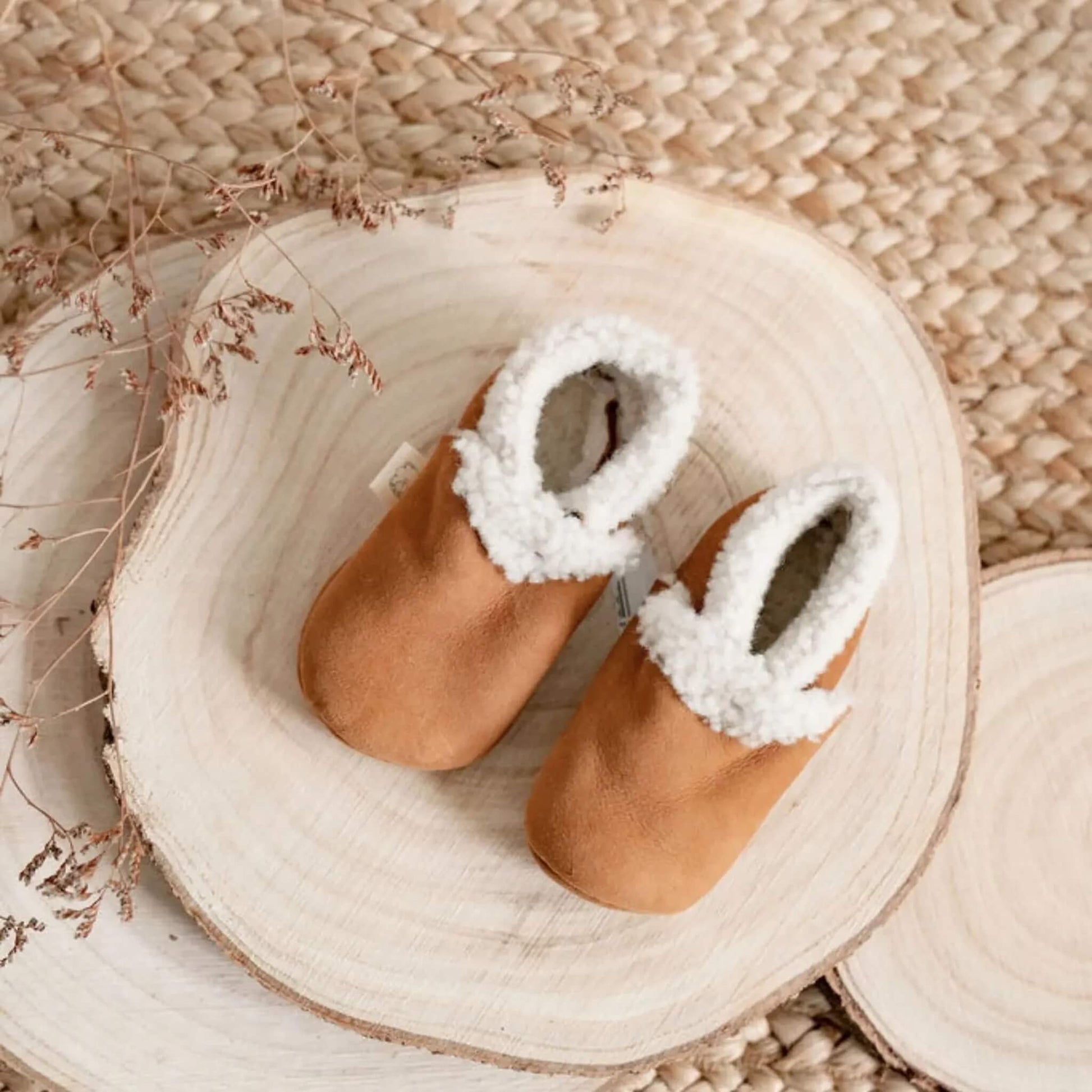 Top view of Baa Baby lambskin booties in tan on a wooden slice, styled with dried florals on a woven mat.