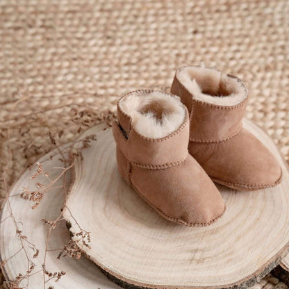 Pair of Baa Baby chestnut sheepskin booties displayed on a wooden slice with dried florals and woven background.