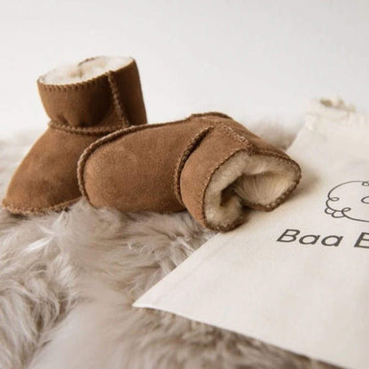 Close-up of Baa Baby chestnut sheepskin booties placed on a fluffy sheepskin rug next to a branded cotton storage bag.