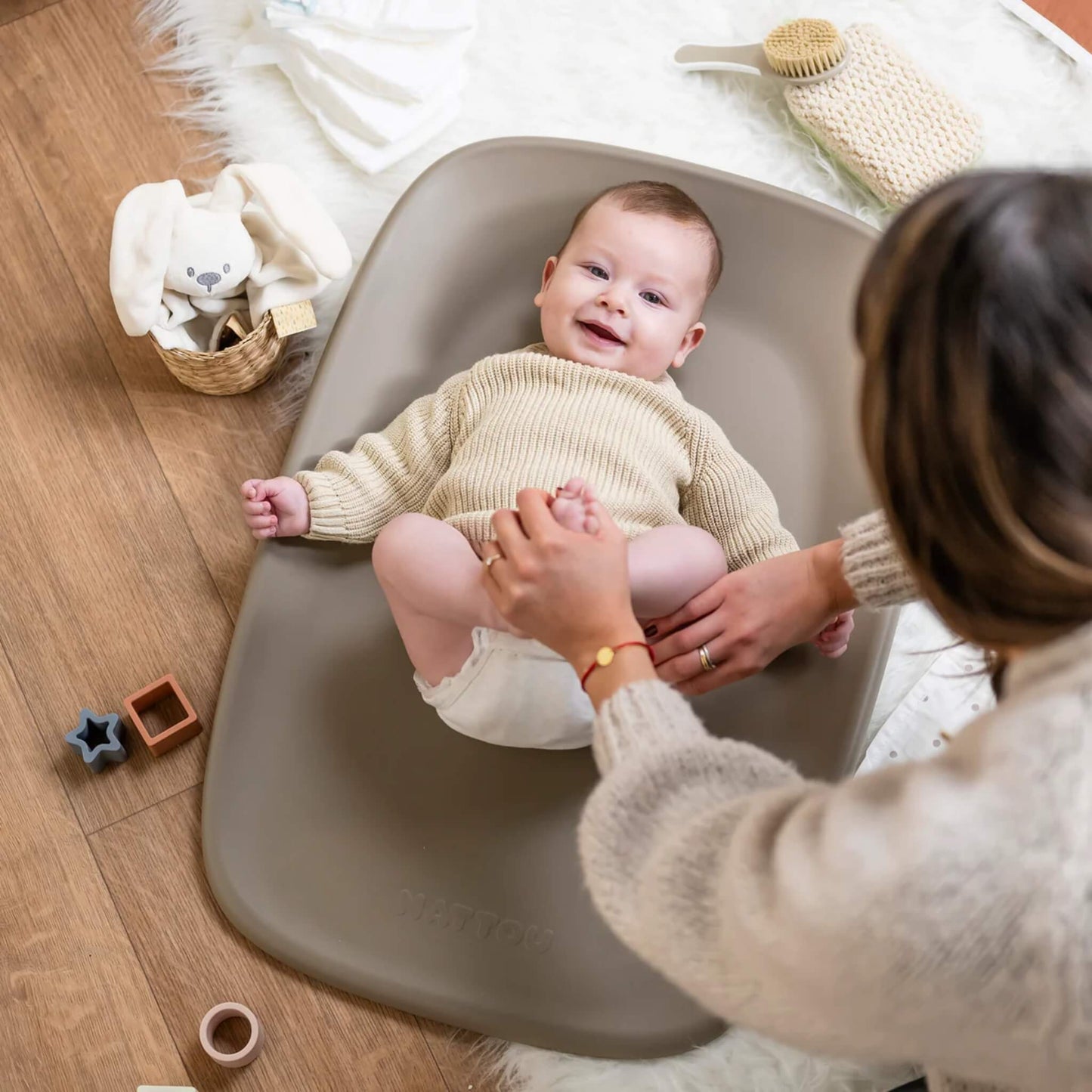 Smiling baby lying on a taupe changing mat while a mother adjusts a nappy, surrounded by soft nursery accessories on a wooden floor.