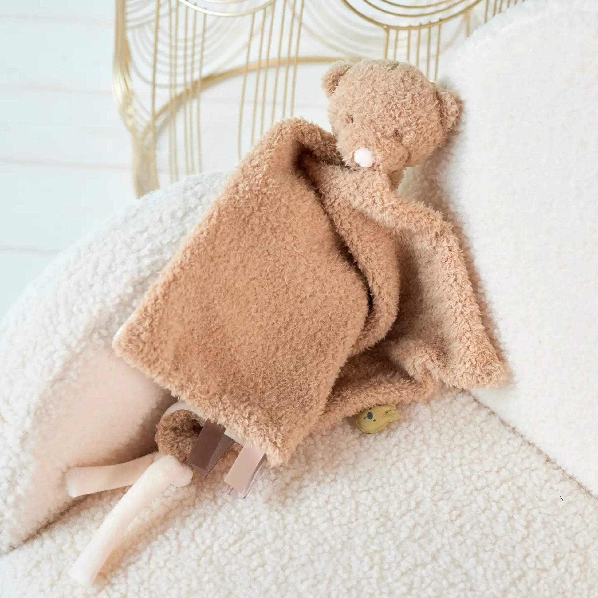Beige teddy bear comforter draped over a soft cream chair, showing textured plush fabric and ribbon tag details.