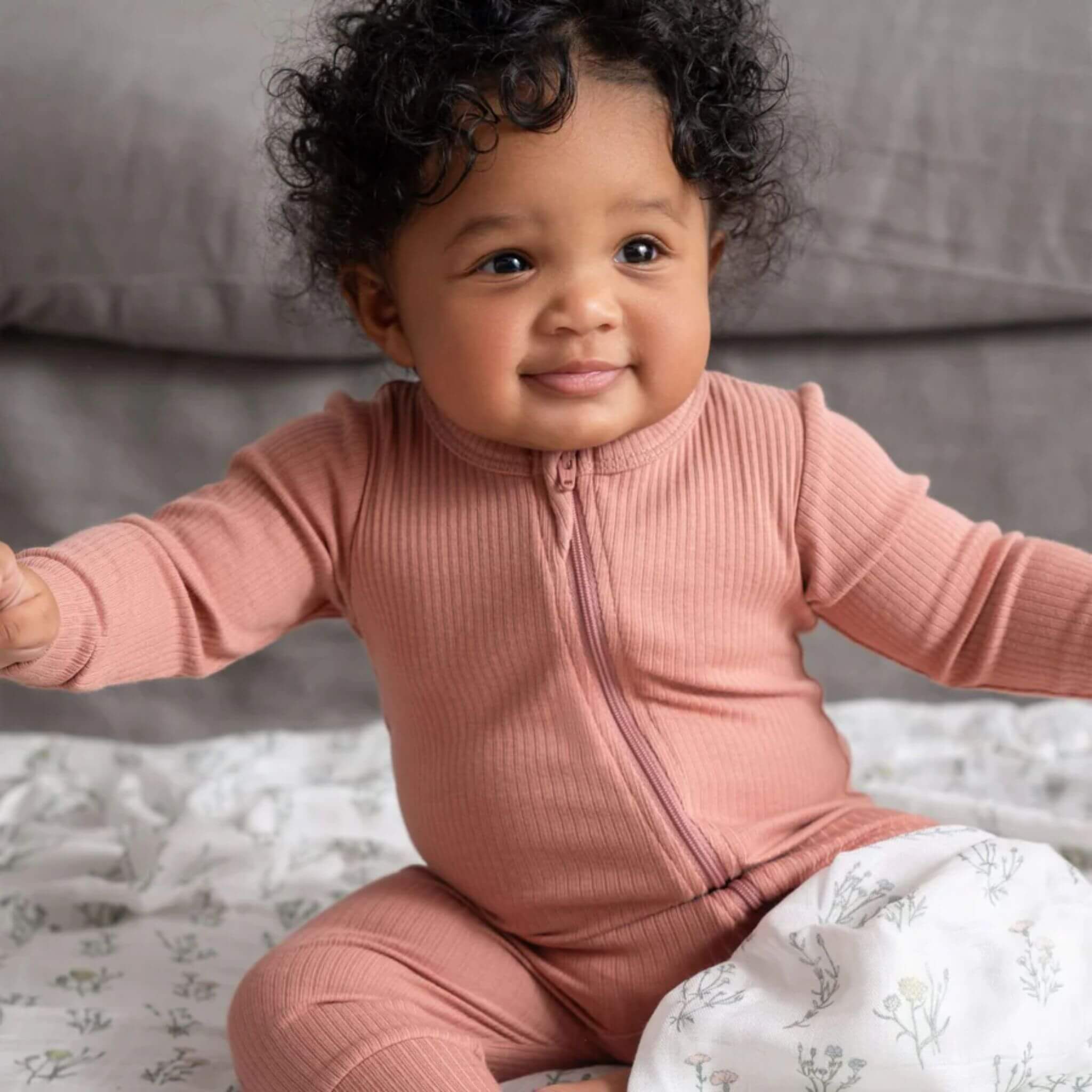 Smiling baby sitting on a soft floral blanket wearing a ribbed dusky rose zip-up sleepsuit. The warm tones and gentle textures create a cosy, natural feel that reflects comfort and quality.