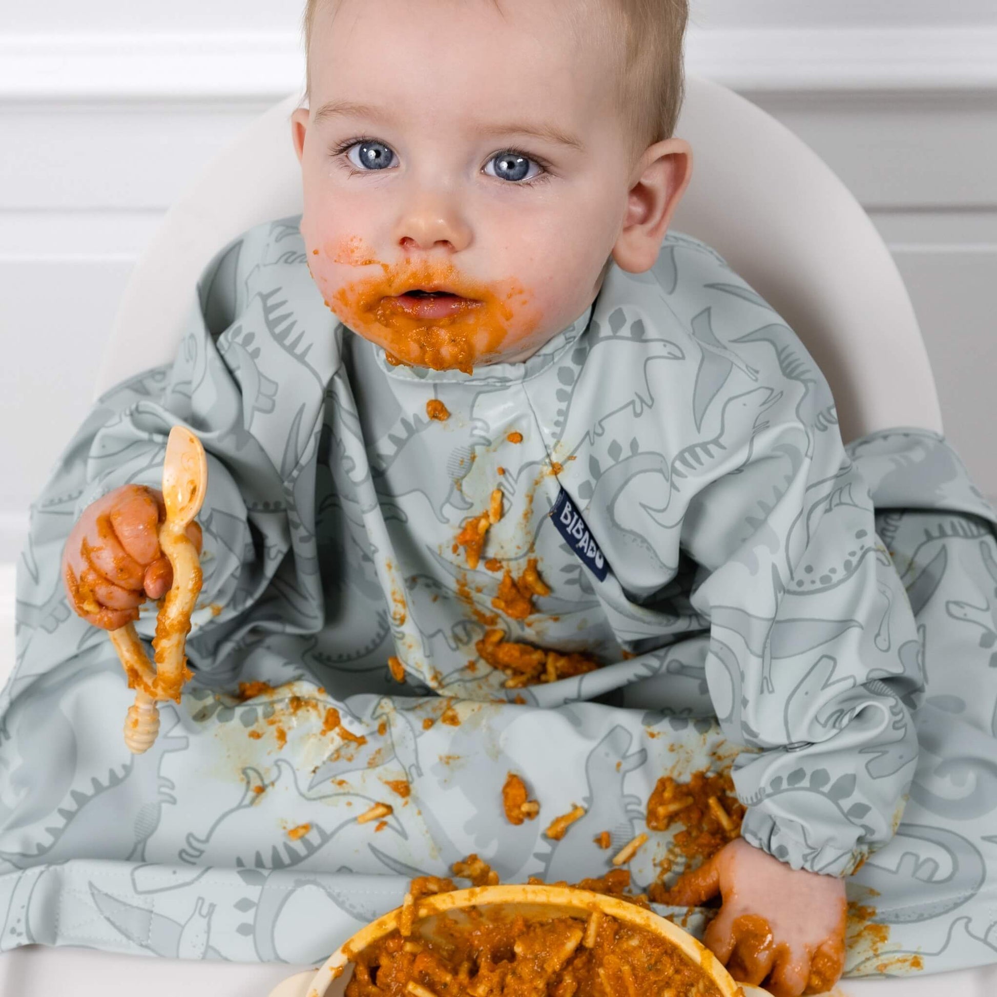 Baby with food-covered face wearing the Dino Roar Bibado bib, showing full coverage and high mess protection.