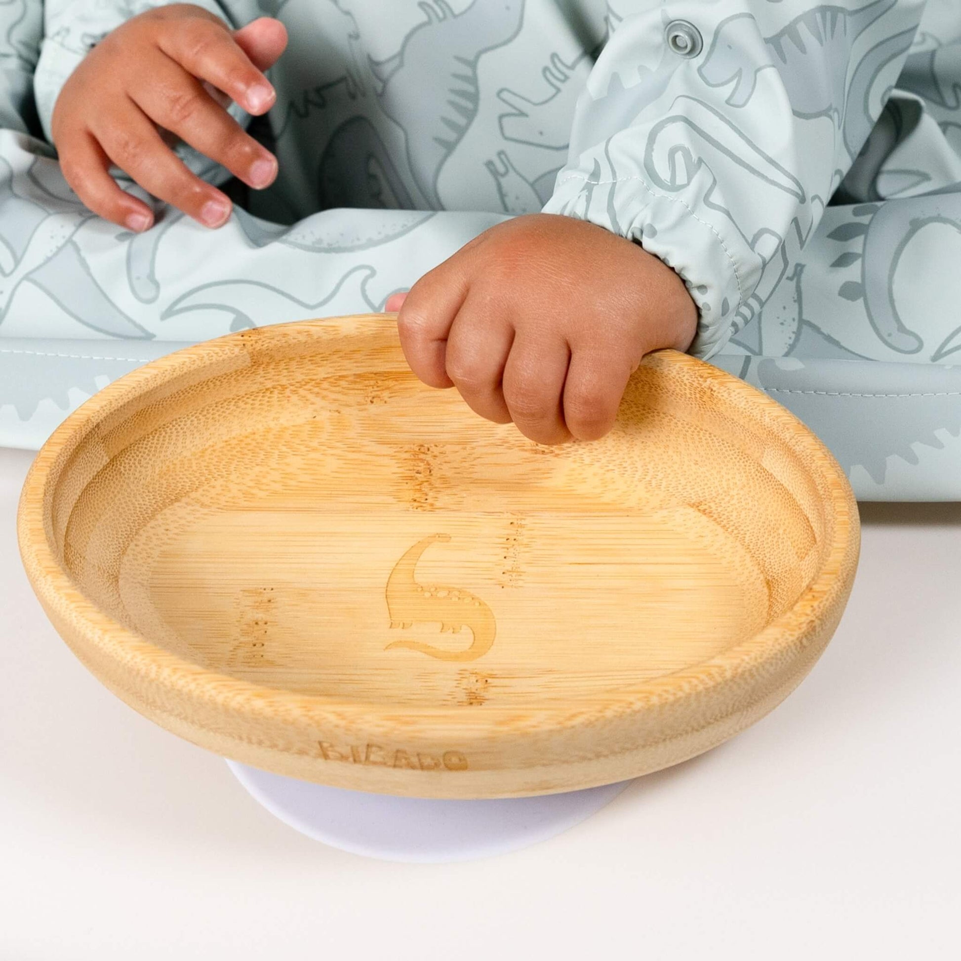 Close-up of a toddler’s hand resting on a wooden suction plate while wearing the dinosaur-print Bibado bib.