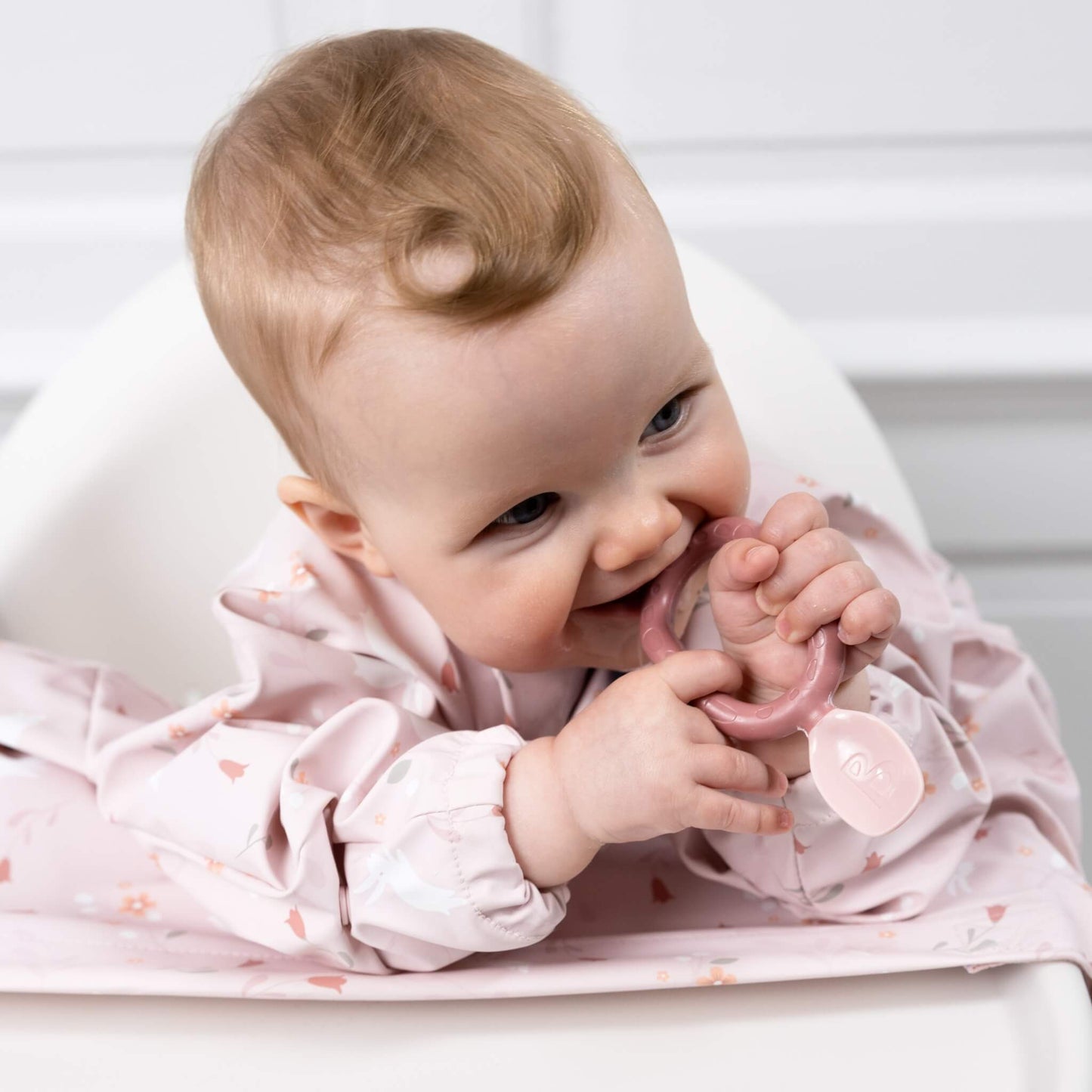 Baby in a highchair chewing on the blush Bibado Dippit spoon, wearing a pink coverall bib with a woodland print.