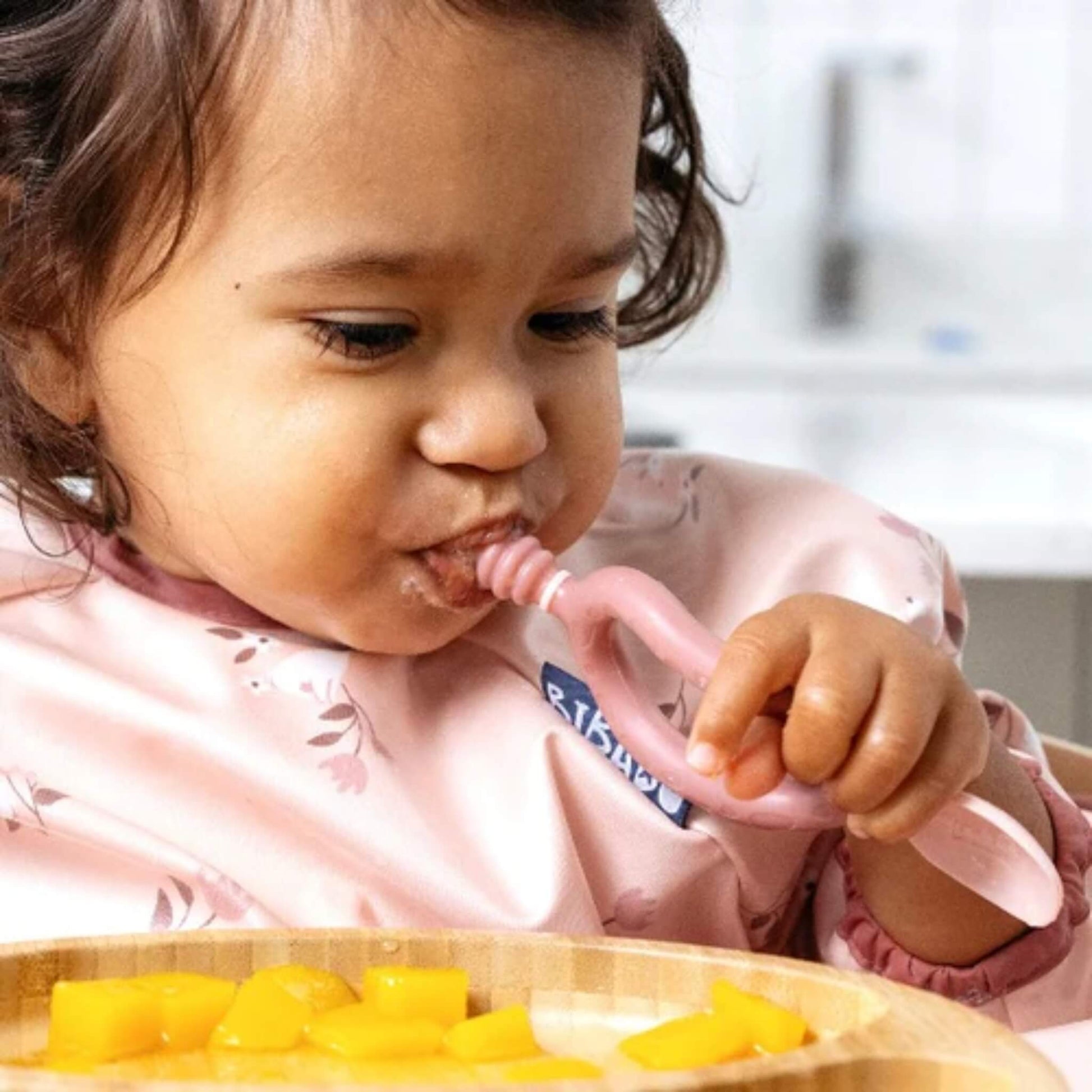 Toddler using the textured dipper end of the Bibado Dippit to self-feed with soft fruit pieces from a bamboo plate.