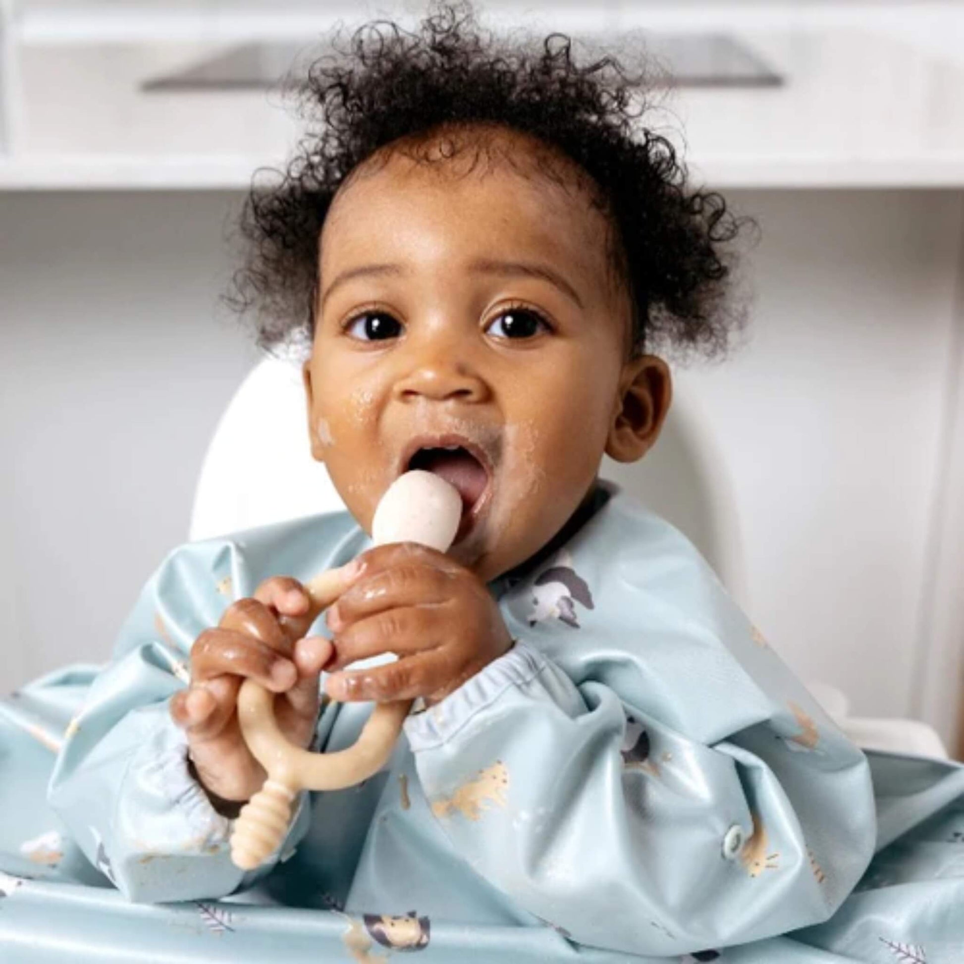 Baby sitting in highchair, wearing a Bibado coverall bib in blue animal print, holding the fawn Bibado Dippit and bringing it to their mouth.