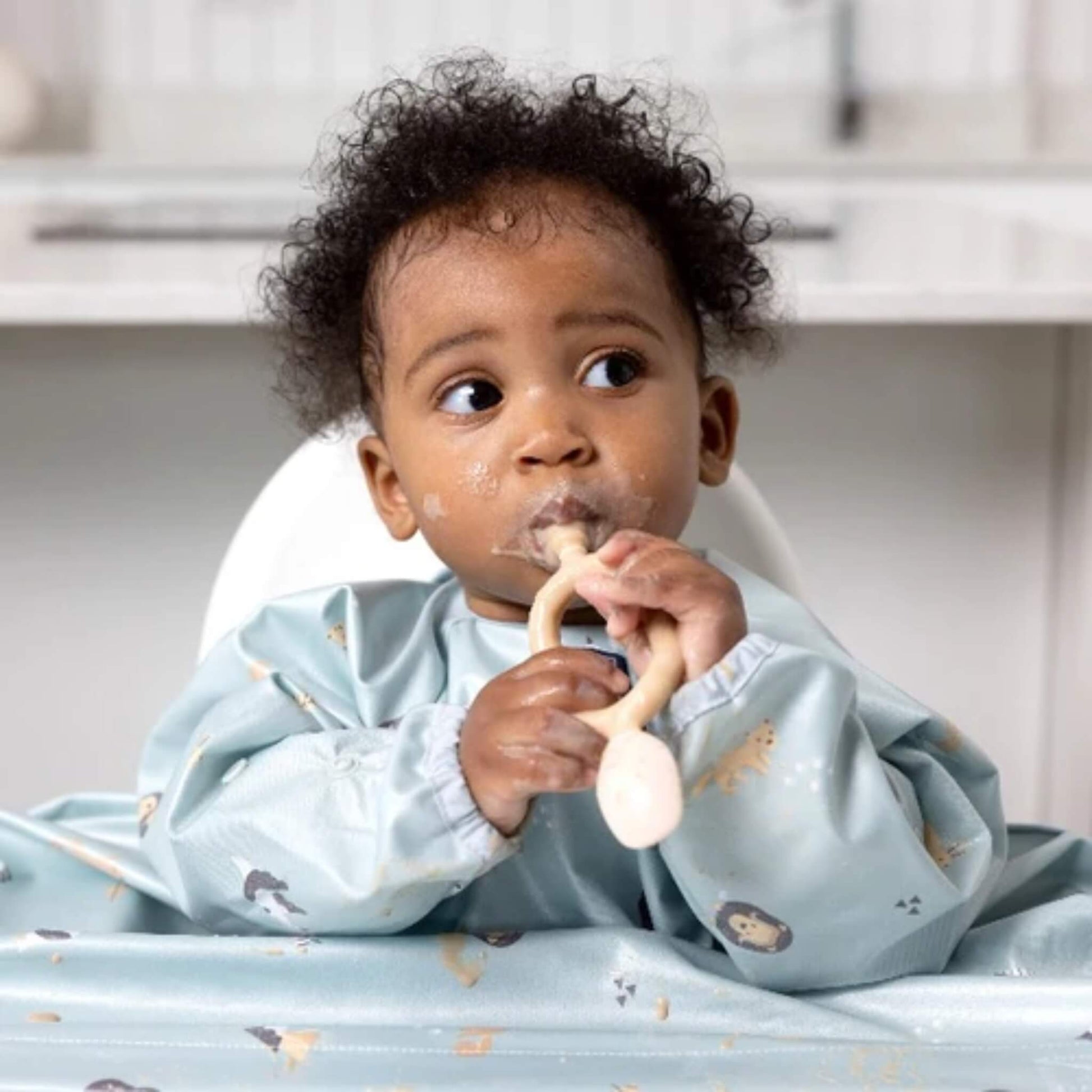 Toddler in a blue animal-print bib seated at a highchair, gripping the fawn Bibado Dippit with both hands while looking to the side.