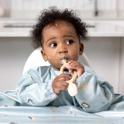 Toddler in a blue animal-print bib seated at a highchair, gripping the fawn Bibado Dippit with both hands while looking to the side.