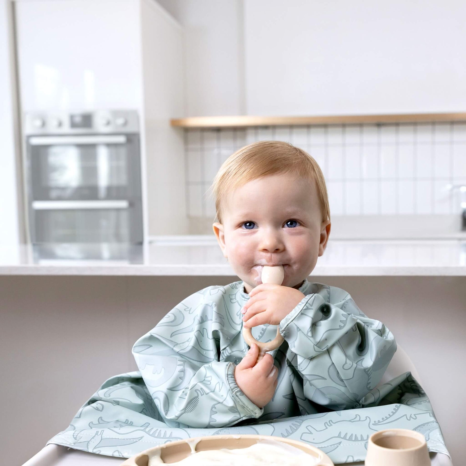 Baby with blonde hair and dinosaur-print bib smiling softly while holding the Bibado Dippit spoon at a kitchen table.
