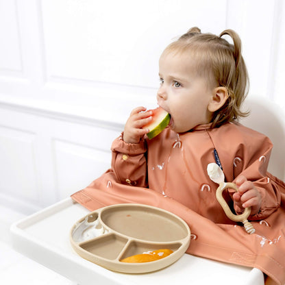 Toddler wearing a chasing rainbows patterned Bibado bib eating watermelon and holding the fawn Bibado Dippit spoon with yoghurt visible on the plate.