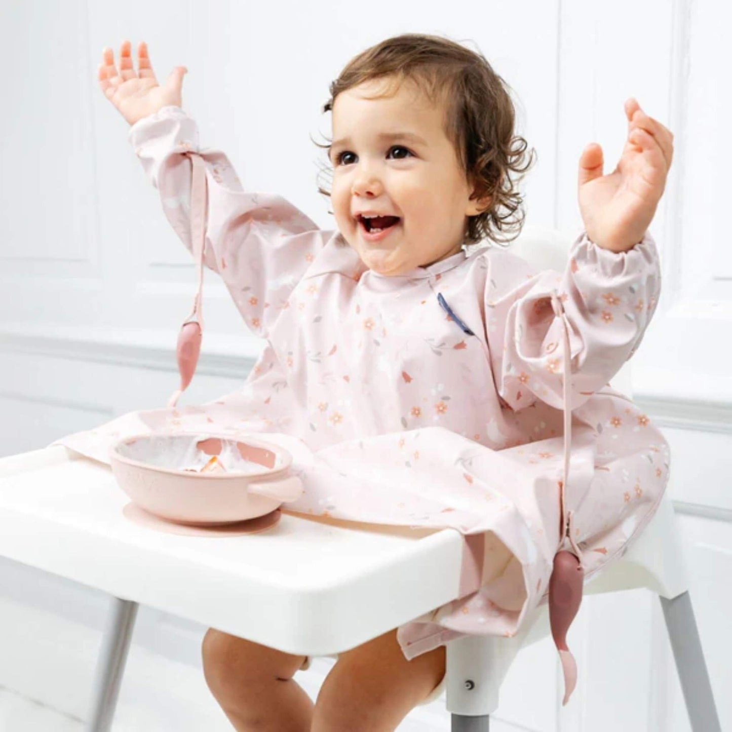 Toddler smiling with arms raised in a highchair, with weaning cutlery safely dangling from their bib straps.