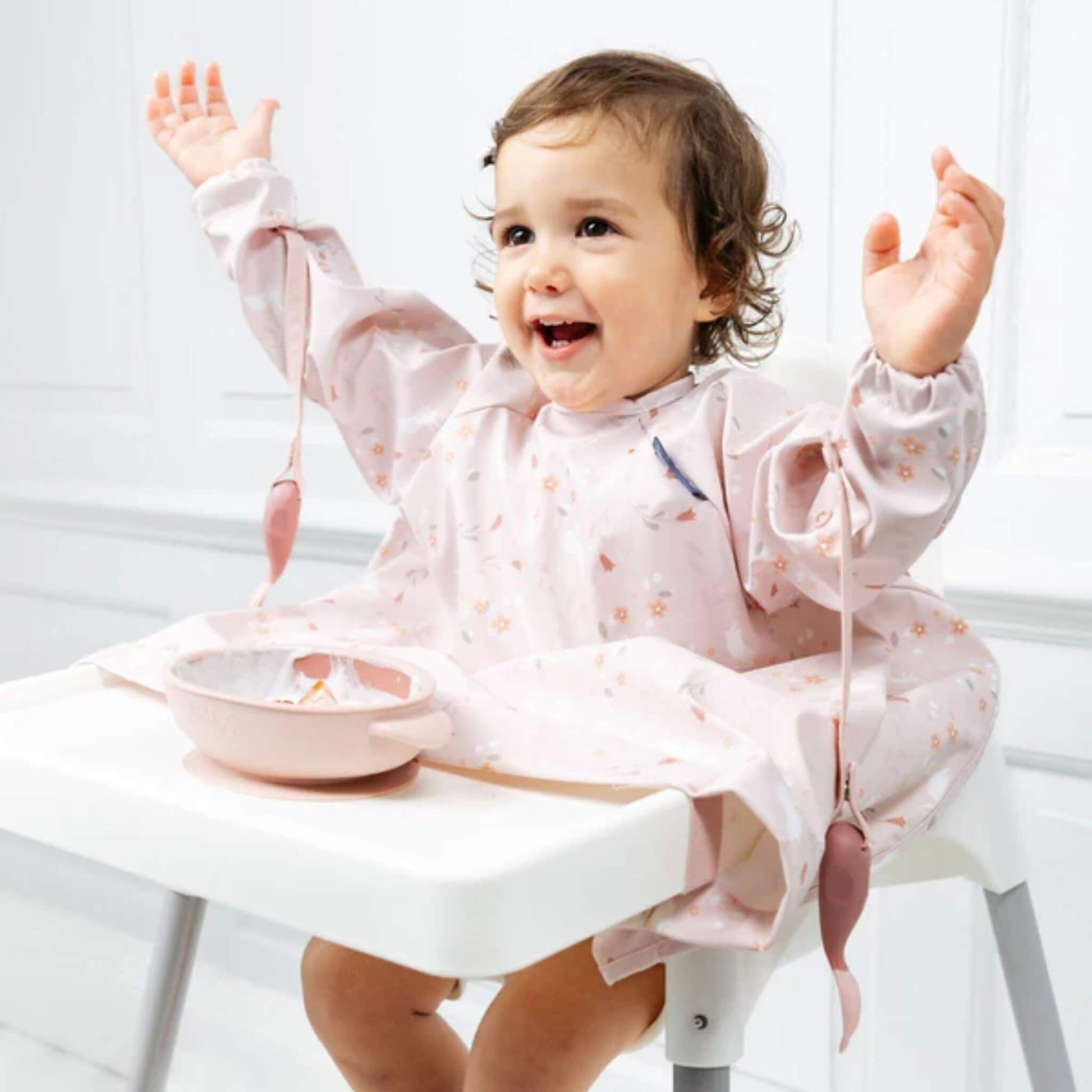 Toddler smiling with arms raised in a highchair, with weaning cutlery safely dangling from their bib straps.