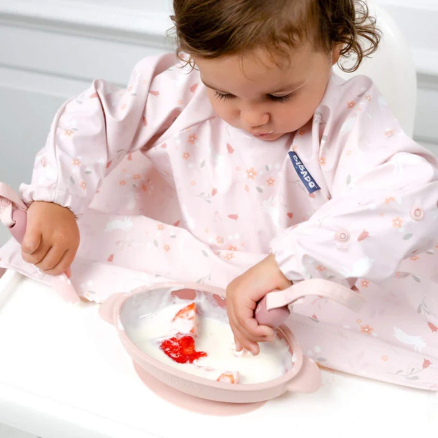Close-up of toddler concentrating while scooping strawberries and yogurt with pink training cutlery, wearing a long-sleeved weaning bib.