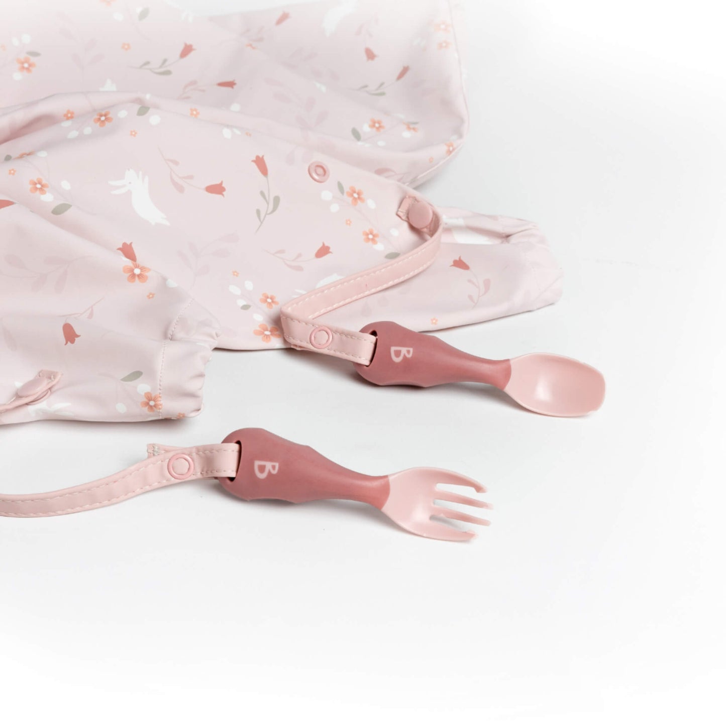 Soft pink spoon and fork clipped to a floral weaning bib, laid flat on a marble surface for prep.