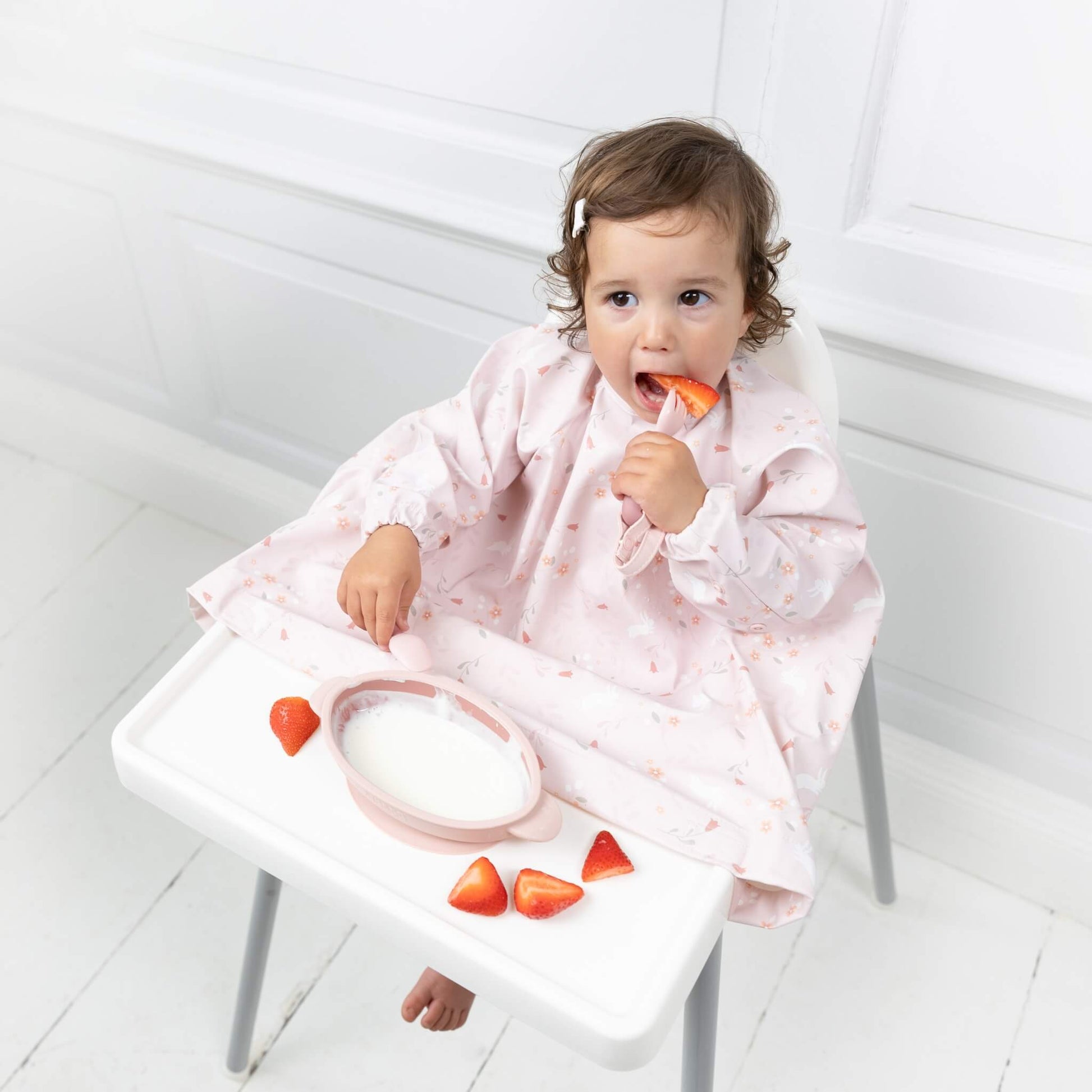 Toddler learning to self-feed using a pink fork to pick up sliced strawberry from a highchair tray.