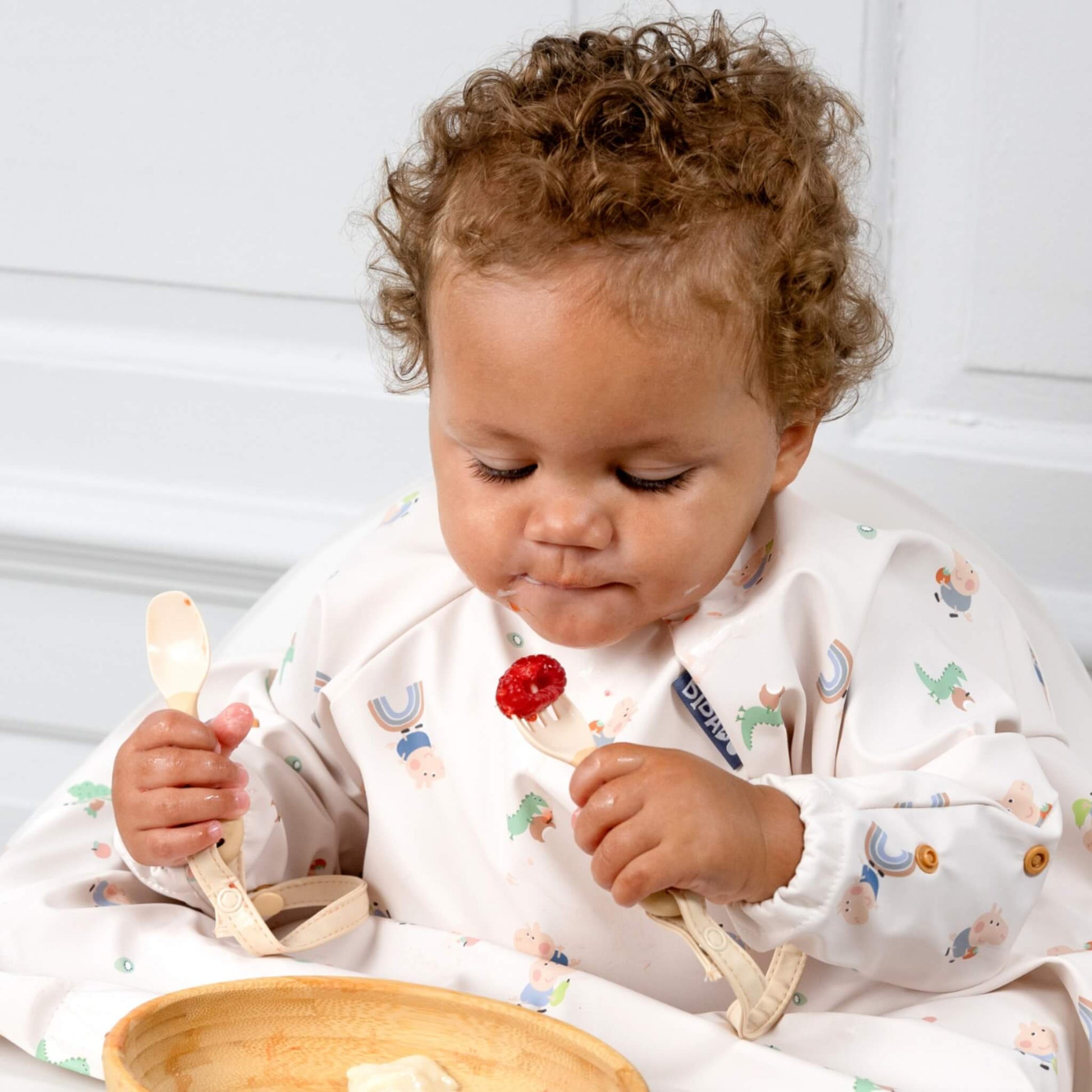 Child practising self-feeding with a raspberry on the fork, wearing a bib with soft animal and rainbow illustrations.