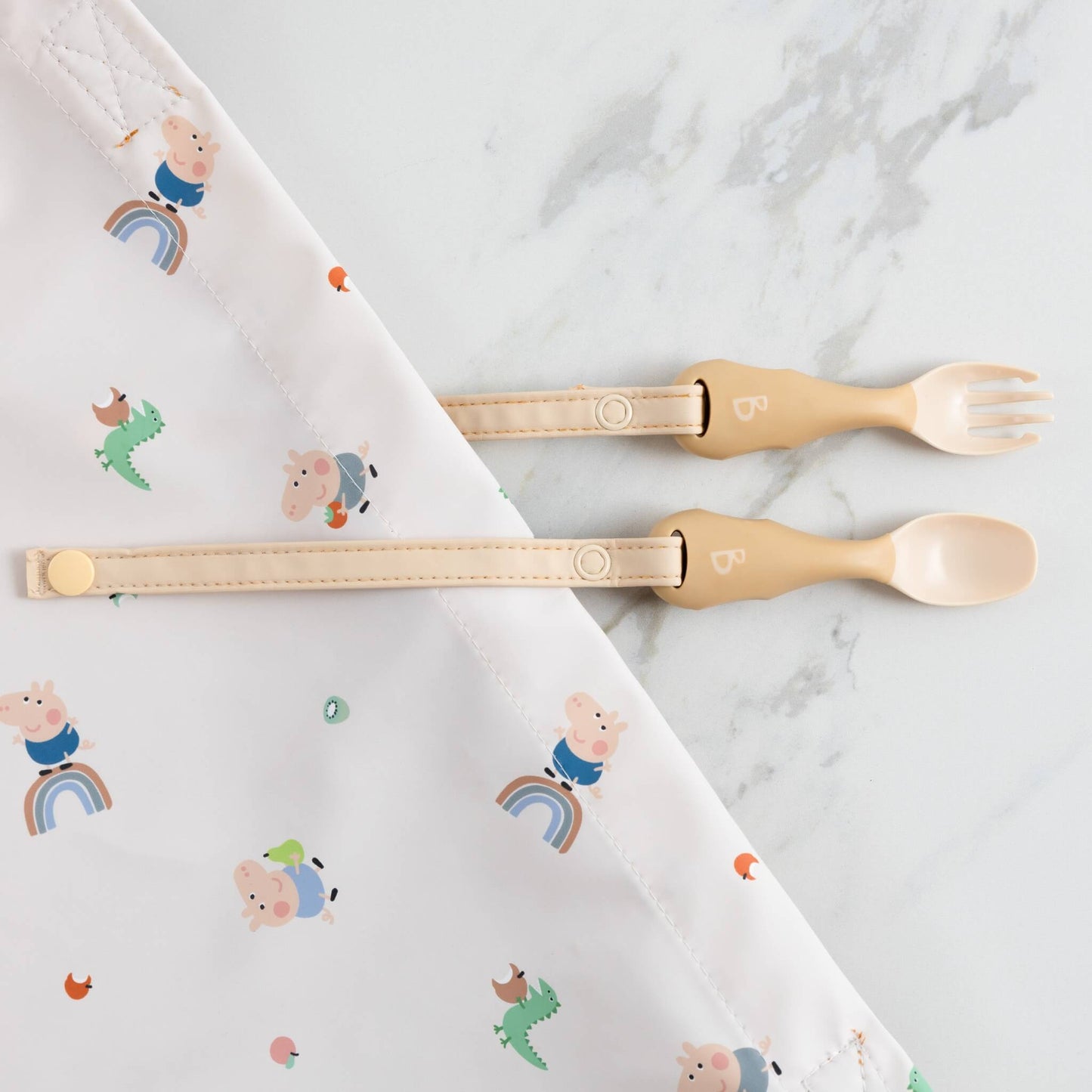 Beige spoon and fork clipped to a white bib with rainbow and Peppa Pig print, positioned on a marble background.