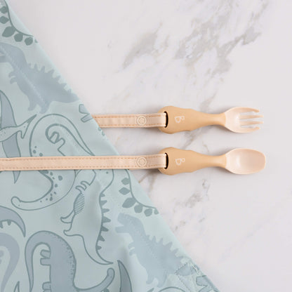 Cutlery set secured to a dinosaur-print bib, popper straps extended across the surface in a feeding-ready setup.