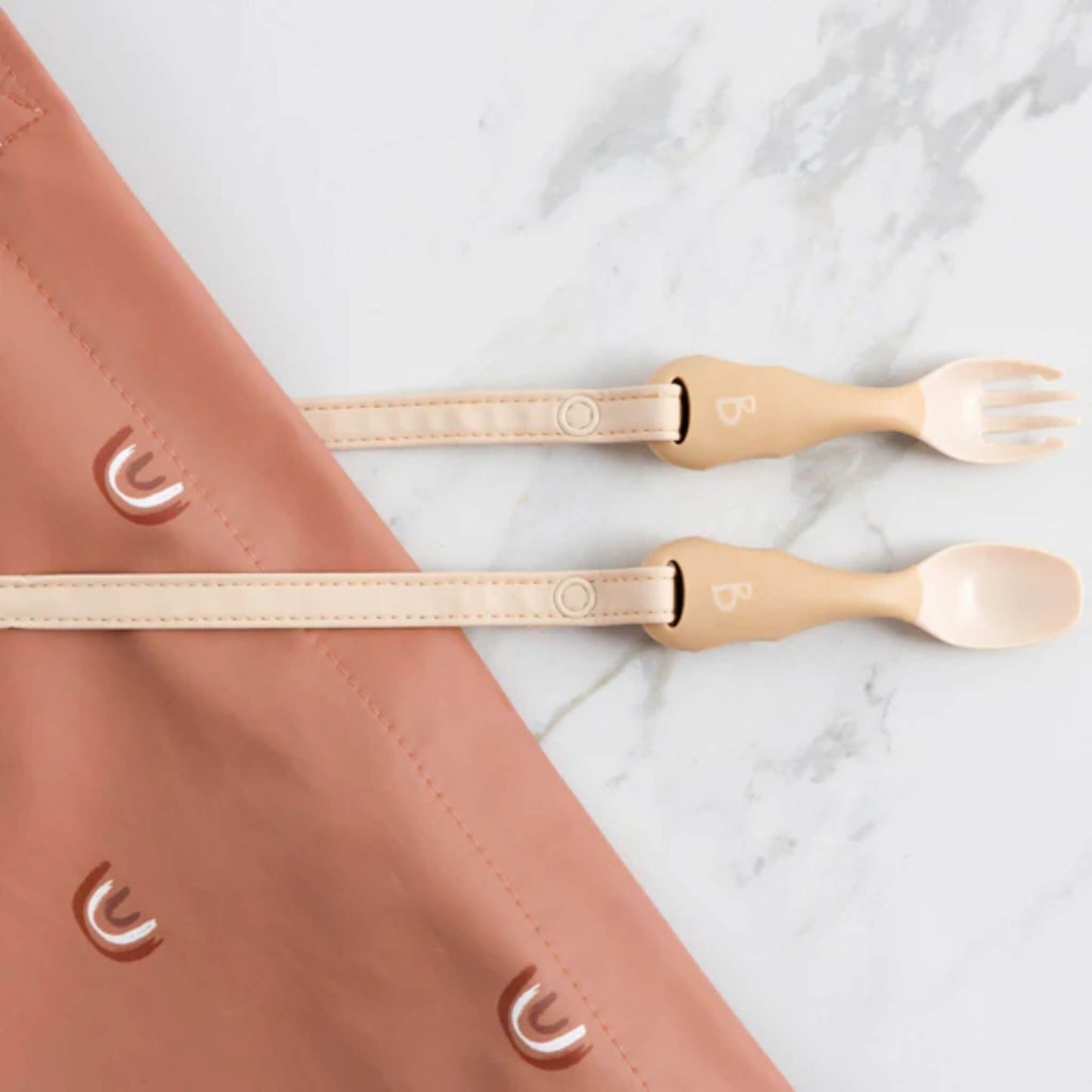 Weaning spoon and fork in soft beige attached to a terracotta bib with white rainbow motifs, laid out for use.