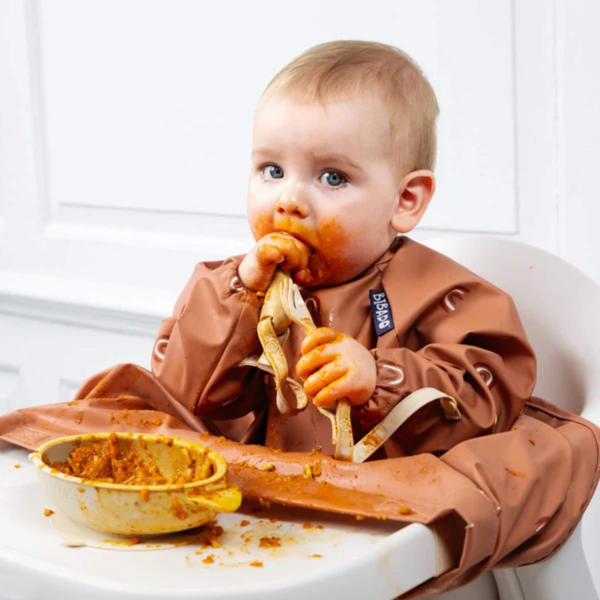 Baby covered in food holding the spoon and fork by the straps after a messy but successful self-feeding session.
