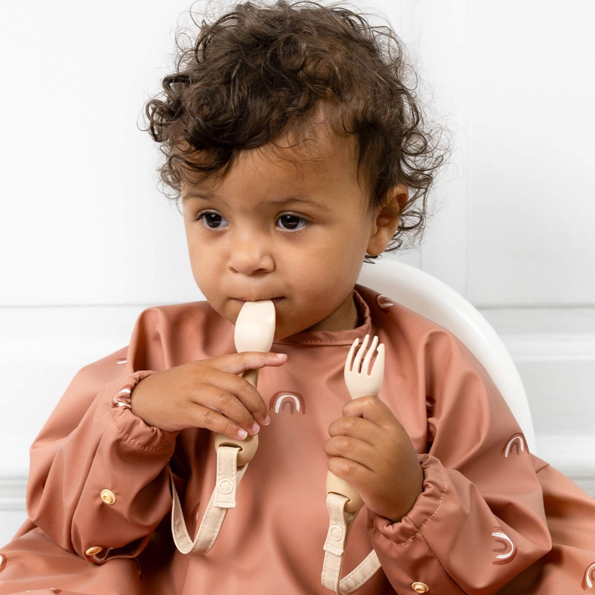 Toddler seated with one utensil in their mouth and the other in hand, straps still clipped to their coverall bib.