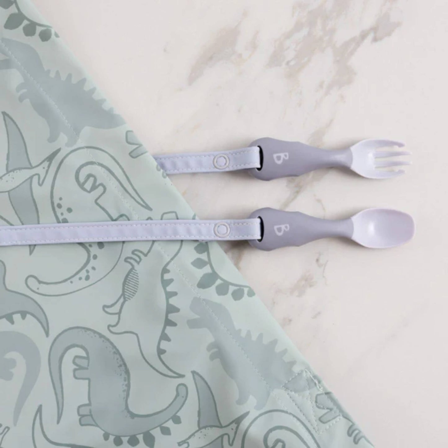 Mist-coloured toddler cutlery clipped to a dinosaur-print bib, laid out on a marble surface ready for weaning.