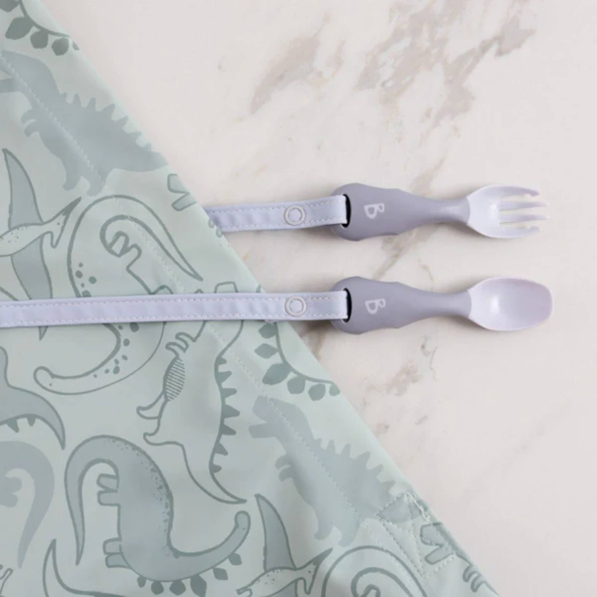 Mist-coloured toddler cutlery clipped to a dinosaur-print bib, laid out on a marble surface ready for weaning.