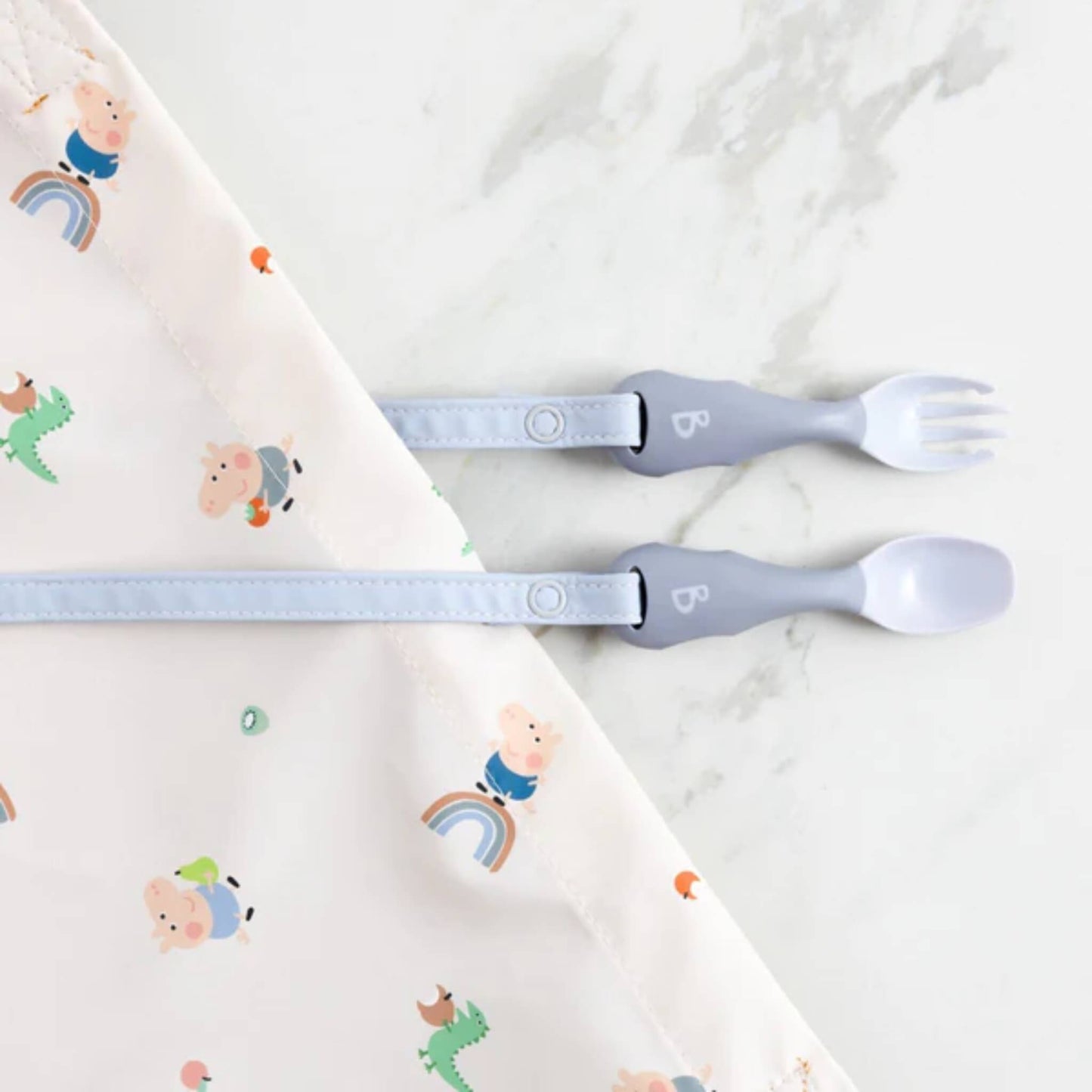 Close-up of the spoon and fork attached to a Peppa Pig print bib, resting on a marble surface.