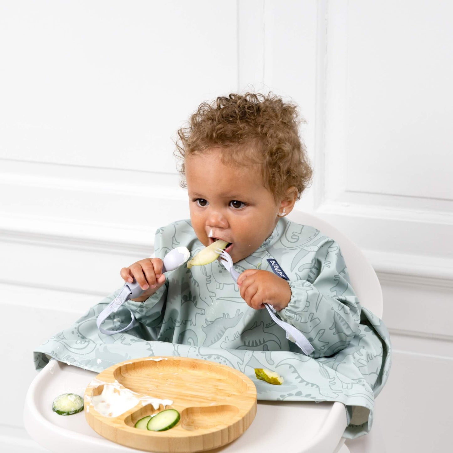 Toddler wearing a dinosaur-print bib using both hands to eat with the Mist cutlery, seated at a highchair.