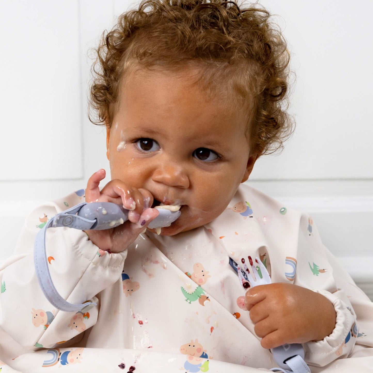 Baby concentrating on self-feeding with food around their mouth, holding both pieces of the Mist cutlery set.