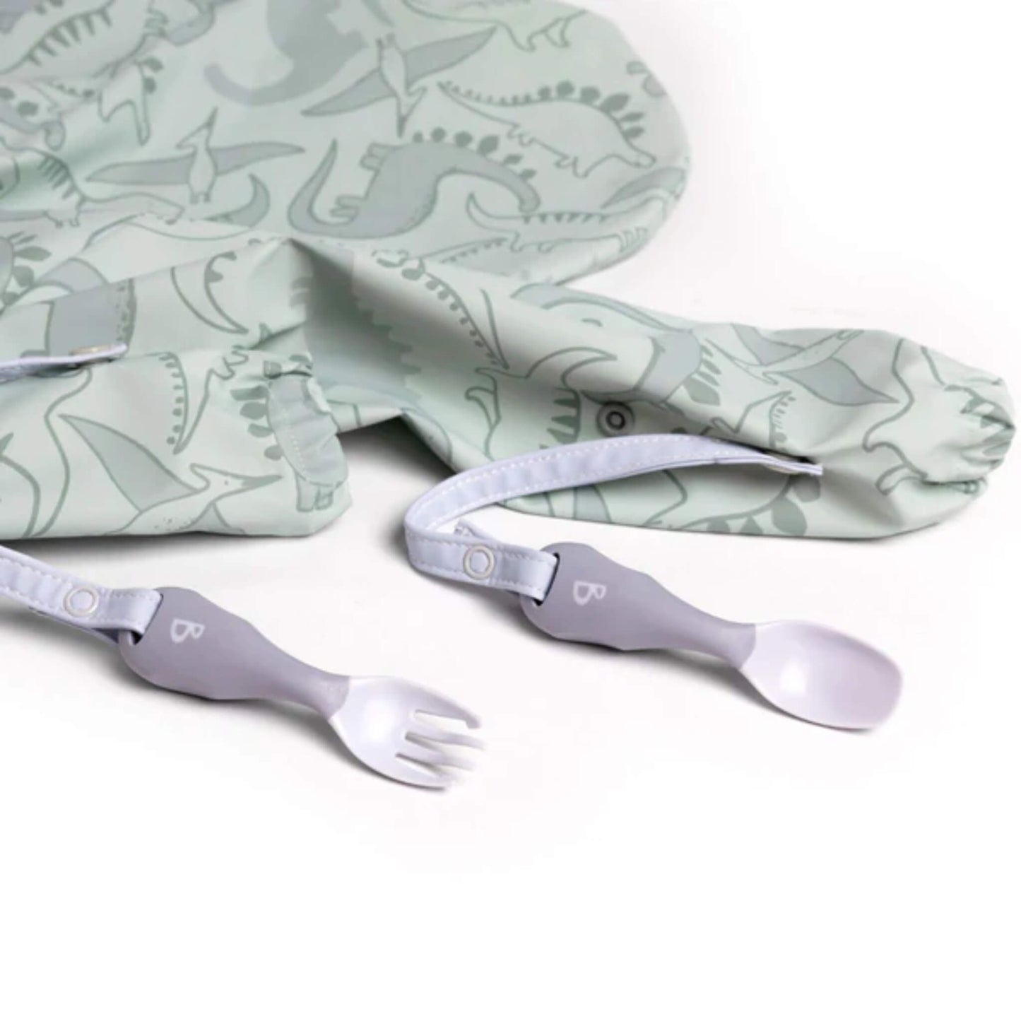 Bibado Handi cutlery laid out beside a dinosaur coverall bib, straps stretched across the surface.
