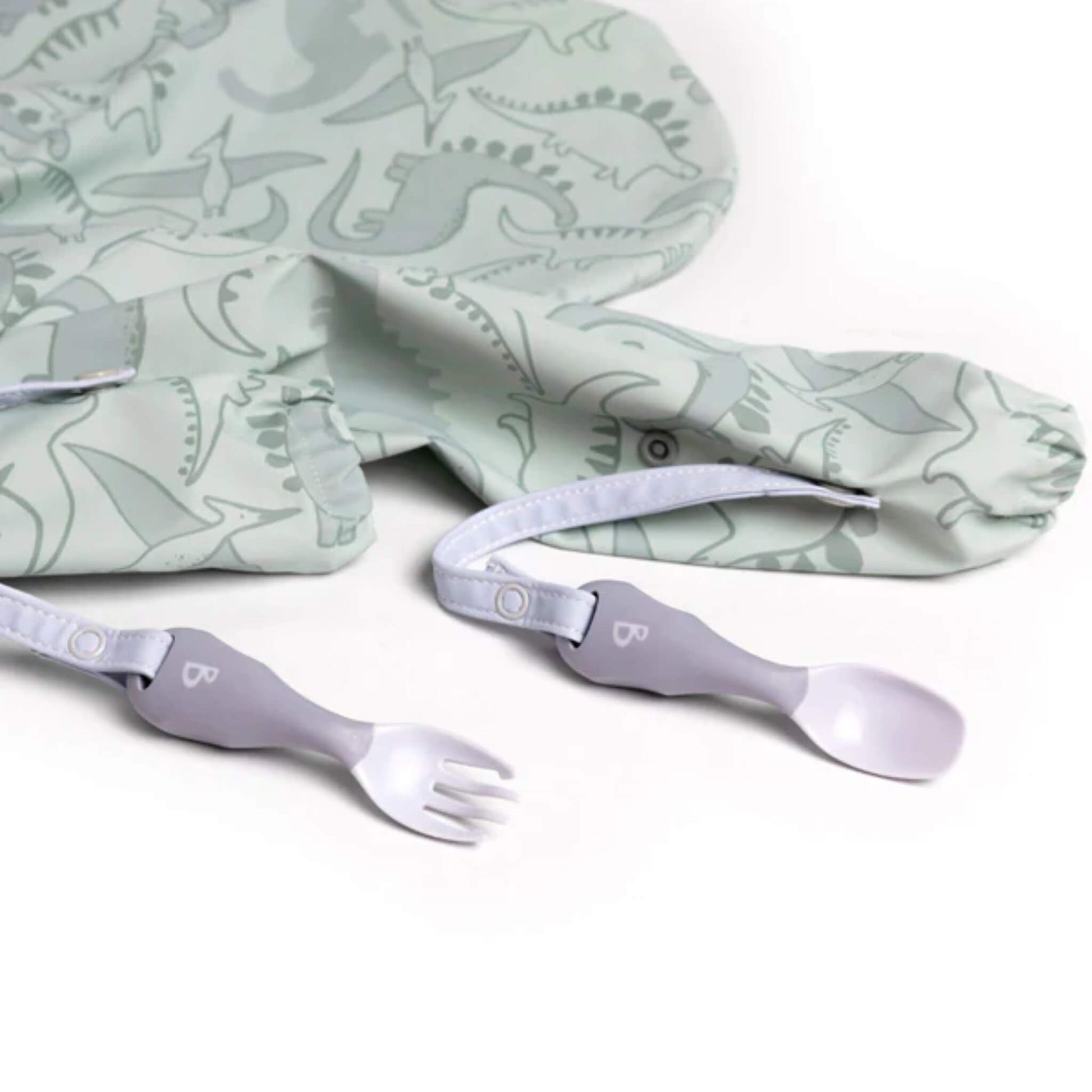 Bibado Handi cutlery laid out beside a dinosaur coverall bib, straps stretched across the surface.