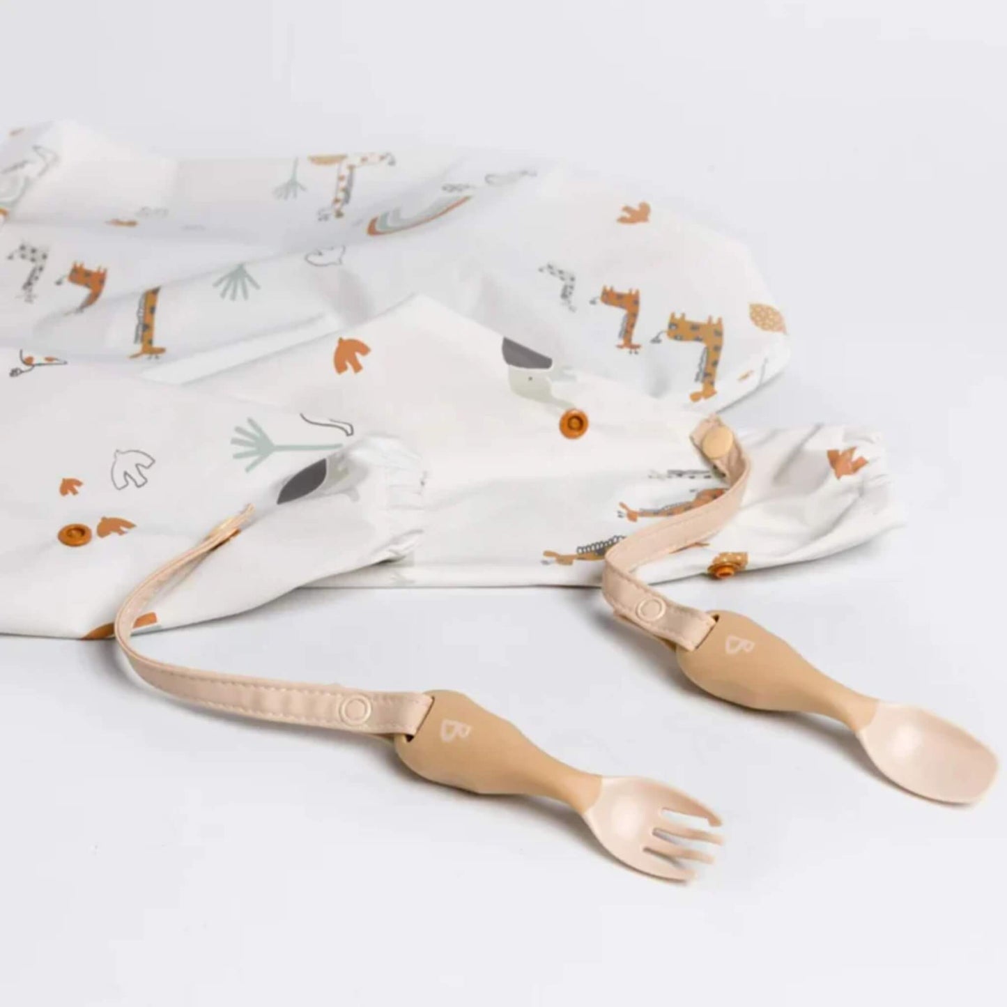 Attachable fork and spoon connected to a Serengeti Spaghetti print coverall bib by adjustable straps on a white surface.