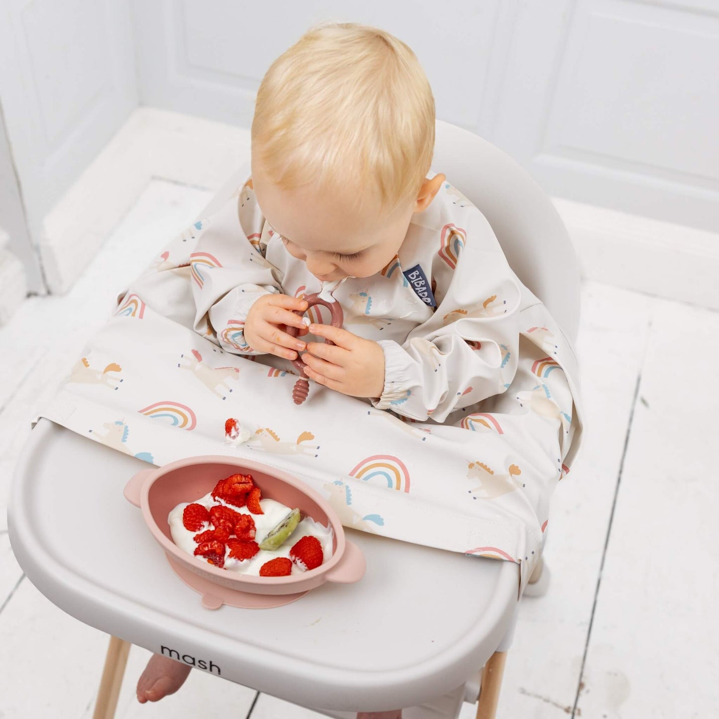 A baby using a blush pink self-feeding utensil during weaning while eating yoghurt, raspberries and kiwi in a highchair.