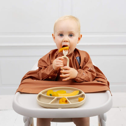 A baby using a fawn self-feeding utensil to eat melon while sitting in a highchair wearing a Bibado coverall bib.