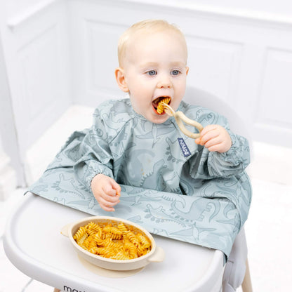 A baby using a fawn self-feeding utensil to scoop and eat pasta independently while wearing a dinosaur print coverall bib.