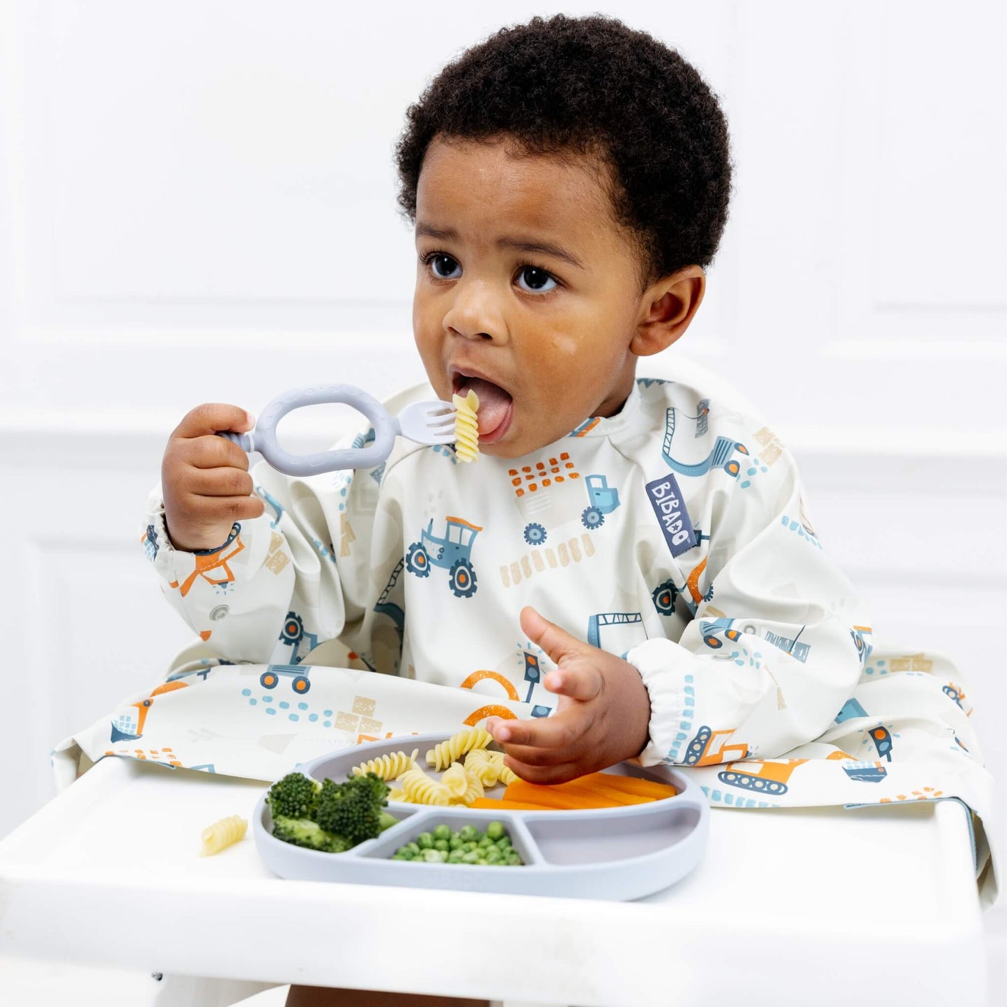 A toddler using a mist blue self-feeding utensil to eat pasta while wearing a construction print coverall bib.