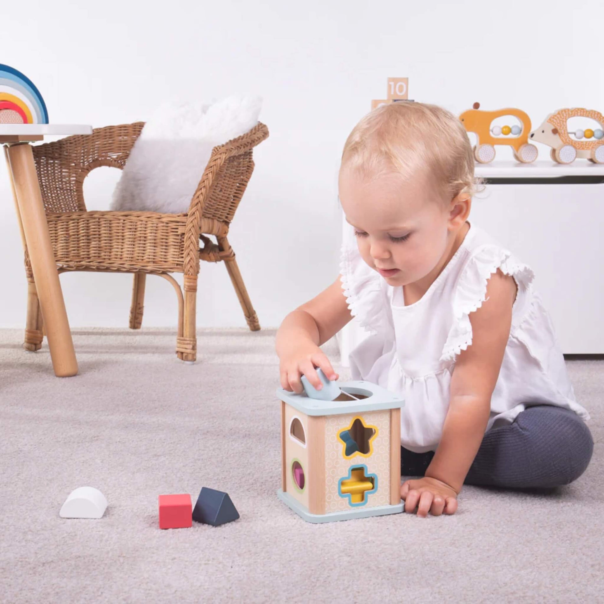 Toddler sitting on the floor placing a shape into the Bigjigs Shape Sorter cube in a nursery setting.