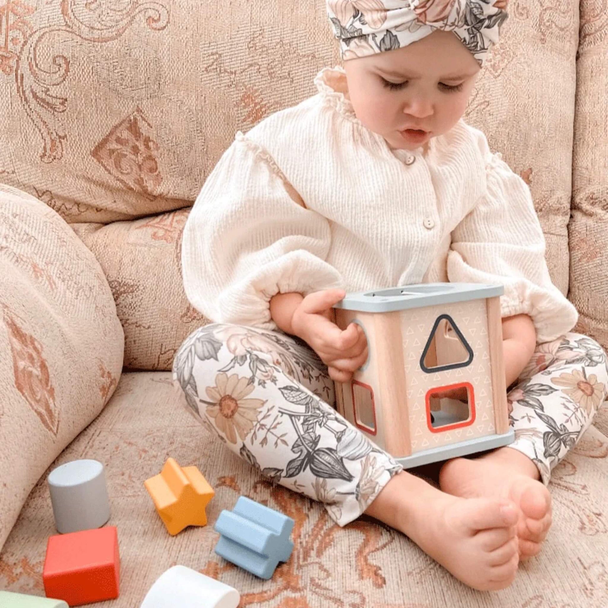 Toddler inspecting the Bigjigs Shape Sorter with assorted shapes laid out on a patterned sofa.