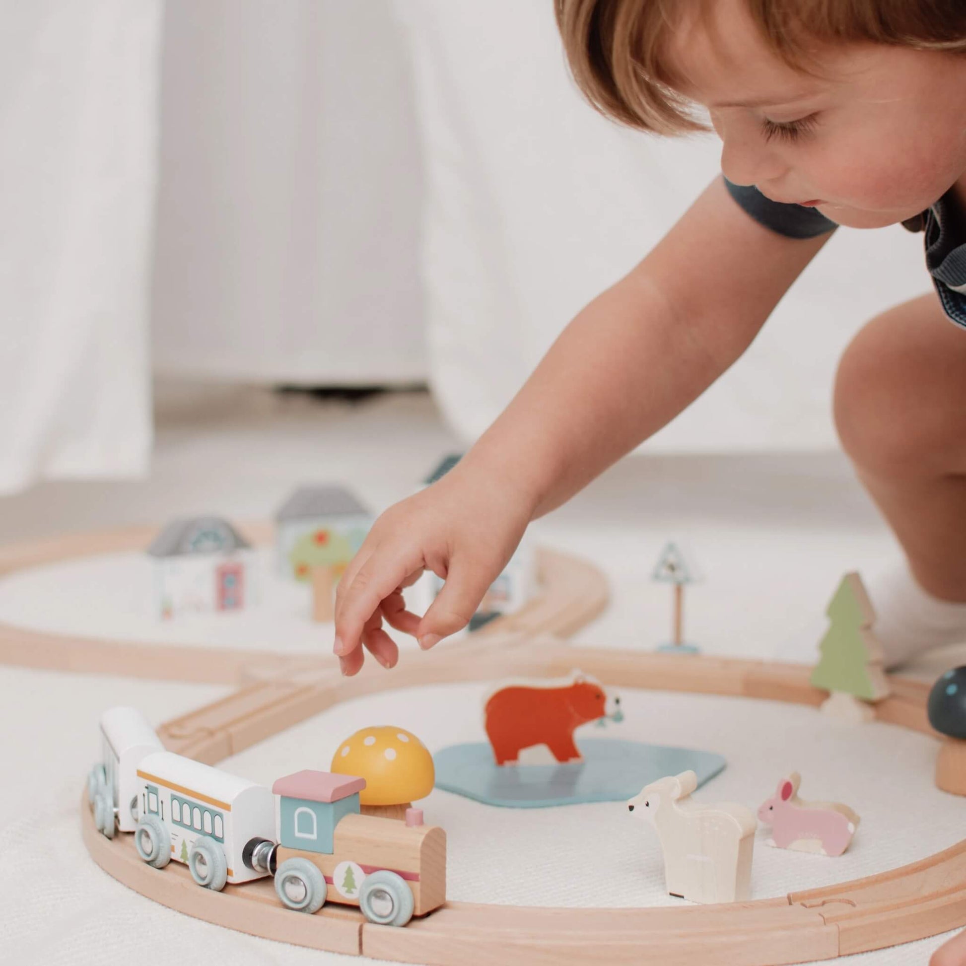 A young child gently placing a train carriage on the wooden track, surrounded by colourful woodland animals and scenery.