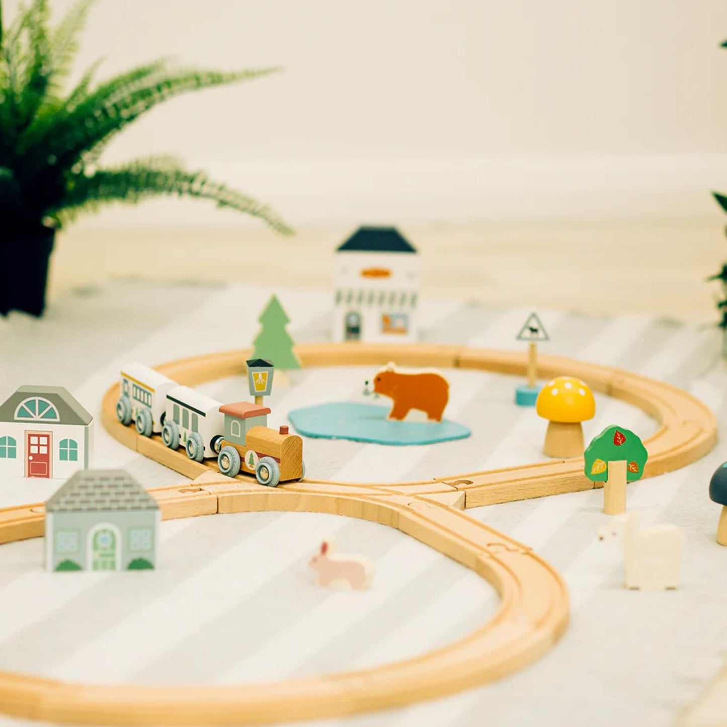 The full train set arranged on a soft striped mat with wooden houses, a bear by the pond, and forest decorations.