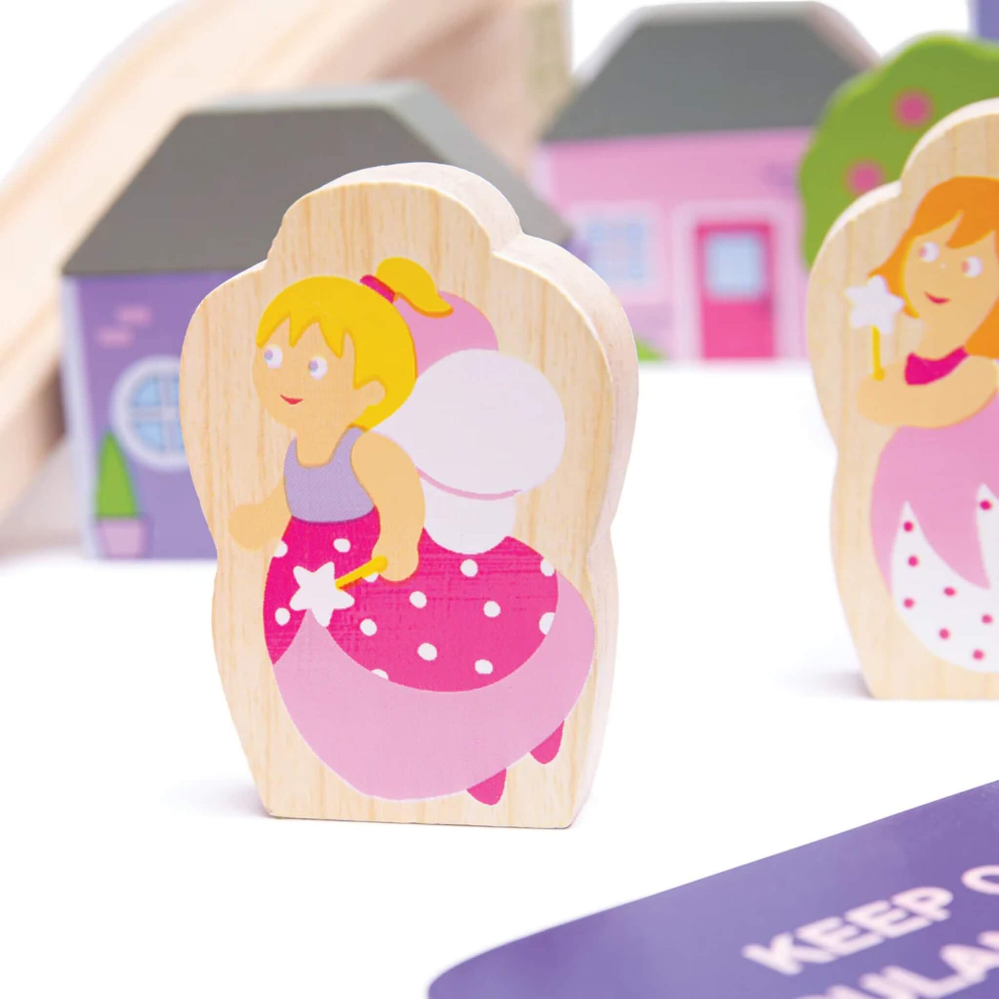 Wooden fairy character with blonde hair, pink wings, and a wand from the Bigjigs Fairy Town Train Set, positioned in front of pastel houses.
