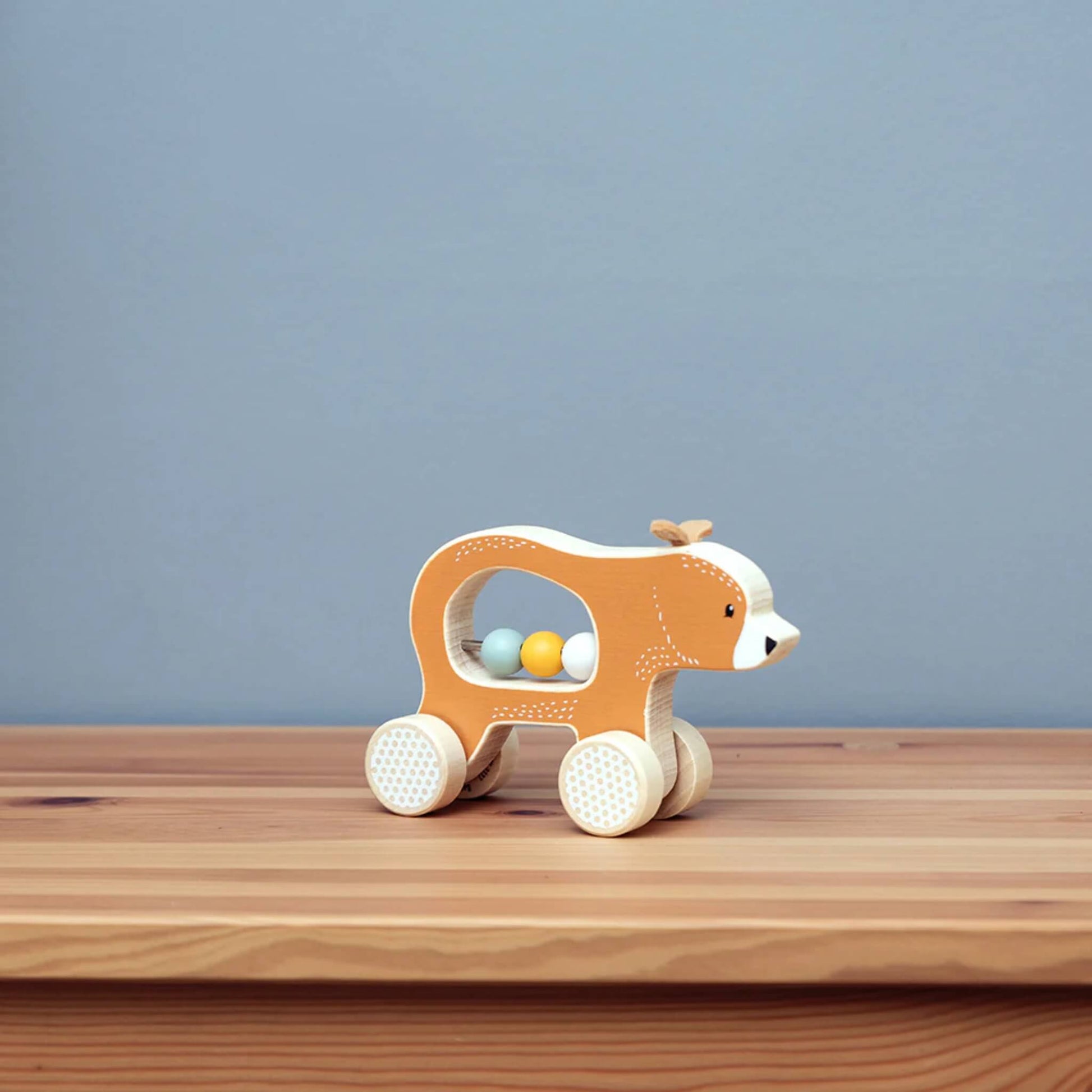 The Push Along Bear toy positioned on a wooden surface with a soft blue background, showing off its smooth shape and natural textures.